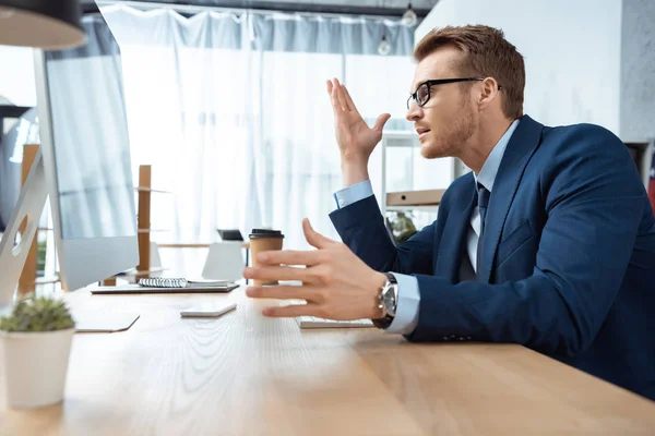 Seitenansicht des gutaussehenden Geschäftsmannes mit Brille gestikuliert mit den Händen am Tisch mit Computermonitor im Büro — Stockfoto