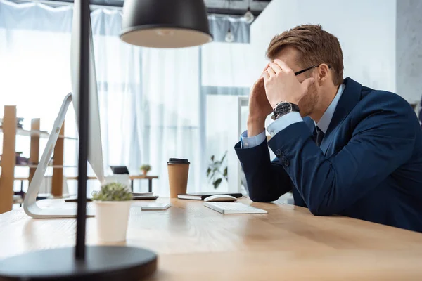 Upset businessman covering face by hands and working at table with computer monitor in modern office — Stock Photo