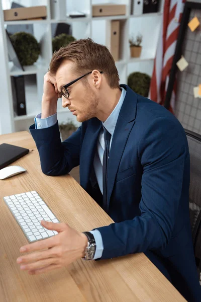 Tired young businessman in eyeglasses gesturing by hand at table with computer keyboard and mouse in office — Stock Photo
