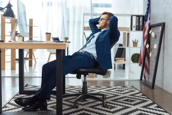 Concentrated businessman in eyeglasses sitting with hands behind head at table with computer monitor in office — Stock Photo