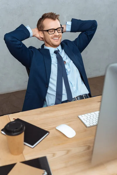 Happy young businessman in eyeglasses sitting with hands behind head at table with computer monitor in office — Stock Photo