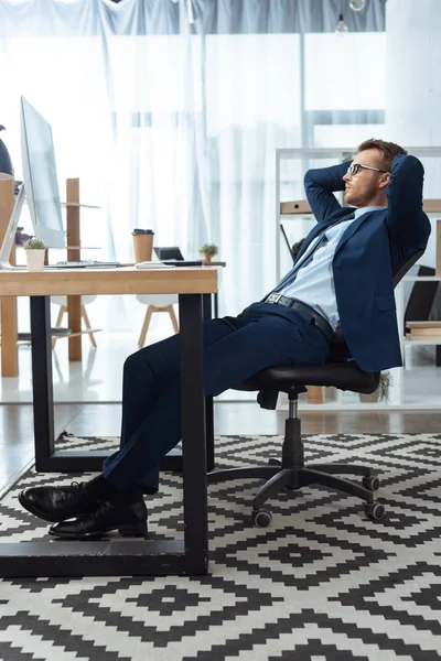 Side view of businessman in eyeglasses sitting with hands behind head at table with computer monitor in office — Stock Photo