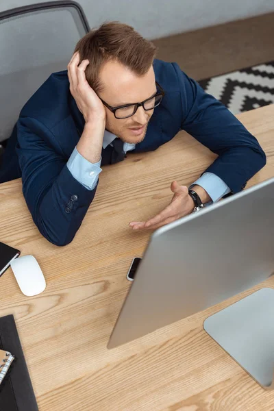 Visão de alto ângulo de empresário irritado em óculos gesticulando à mão na mesa com monitor de computador no escritório — Fotografia de Stock