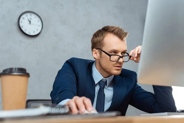 Empresário concentrado ajustando óculos e trabalhando à mesa com monitor de computador no escritório moderno — Fotografia de Stock