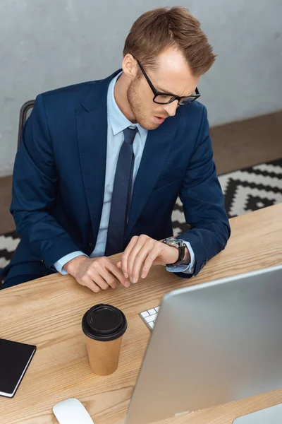 Високий кут зору бізнесмен в окулярах перевіряє годинник за столом з чашкою кави і комп'ютером в офісі — стокове фото