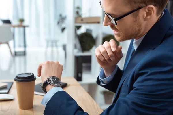 Foco seletivo do empresário emocional em óculos verificando relógio à mesa com xícara de café no escritório — Fotografia de Stock