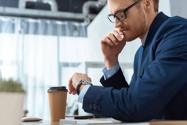 Serio uomo d'affari in occhiali che controlla orologio a tavola con tazza di caffè di carta in ufficio — Foto stock