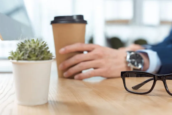 Abgeschnittenes Bild eines Geschäftsmannes, der mit Einweg-Kaffeetasse am Tisch mit Topfpflanze und Brille im modernen Büro sitzt — Stockfoto