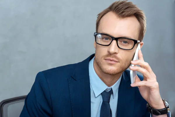 Portrait of handsome businessman in eyeglasses talking on smartphone and looking at camera in office — Stock Photo