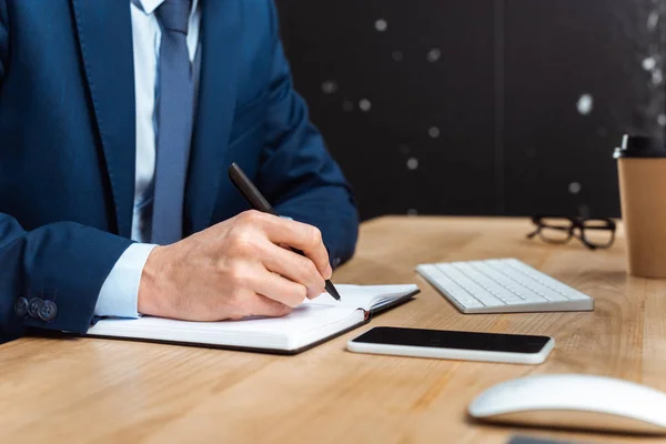 Immagine ritagliata di uomo d'affari che scrive nel libro di testo a tavola con smartphone in ufficio moderno — Foto stock