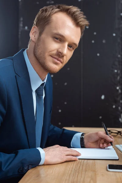 Feliz joven empresario mirando a la cámara y la escritura en el libro de texto en la mesa con el teléfono inteligente en la oficina moderna - foto de stock