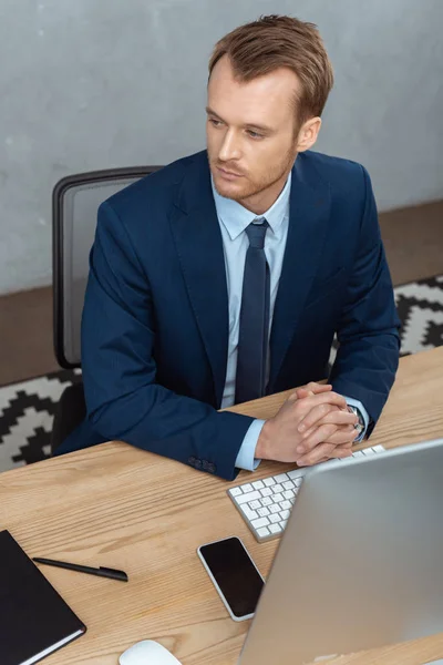 Vista de ángulo alto del hombre de negocios guapo sentado en la mesa con el ordenador y el teléfono inteligente en la oficina moderna - foto de stock