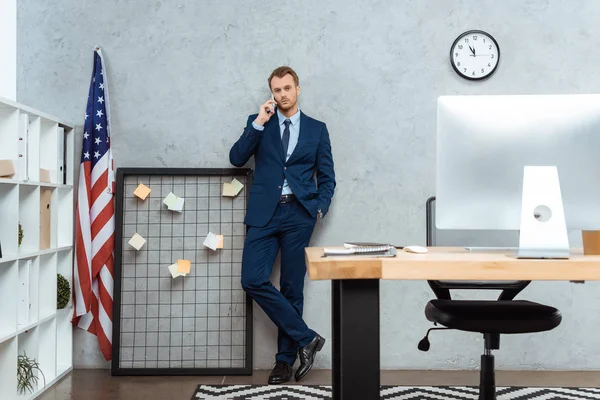 Joven hombre de negocios en traje hablando en el teléfono inteligente cerca de bandera americana por la pared en la oficina moderna - foto de stock
