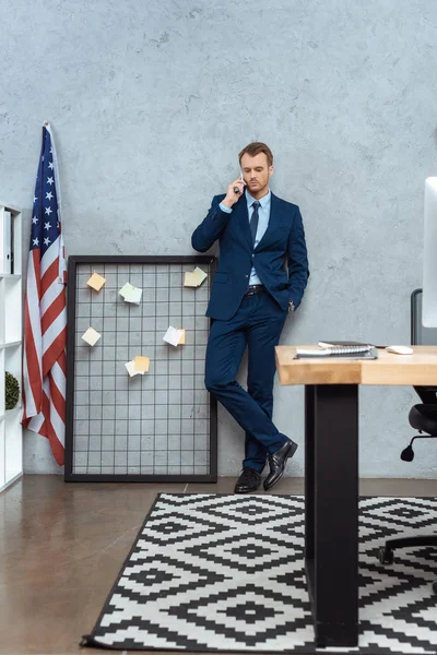 Вибірковий фокус бізнесмена в костюмі, який говорить на смартфоні біля прапора США стіною в сучасному офісі — стокове фото