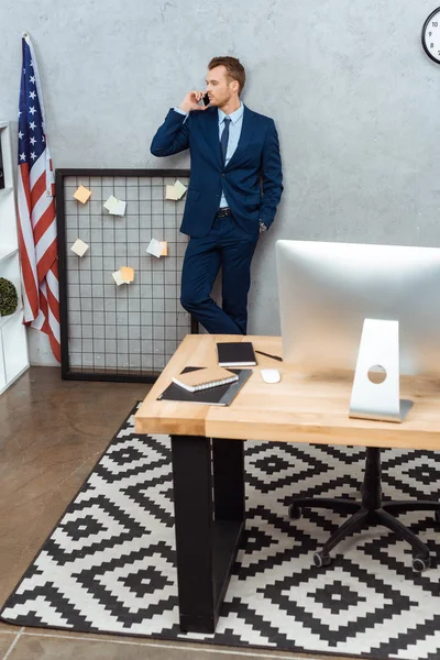 Vista de ángulo alto del hombre de negocios en traje hablando en el teléfono inteligente cerca de bandera americana por la pared en la oficina moderna - foto de stock