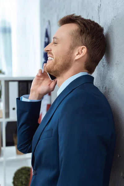 Vue de côté de l'homme d'affaires heureux en costume parler sur smartphone dans le bureau moderne — Photo de stock