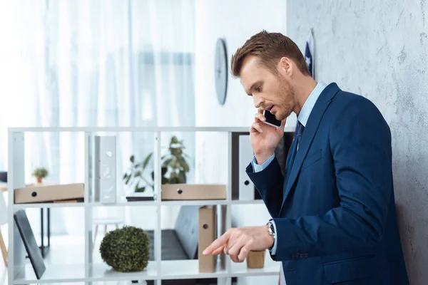 Hombre de negocios serio en traje hablando en el teléfono inteligente y el gesto a mano en la oficina moderna - foto de stock