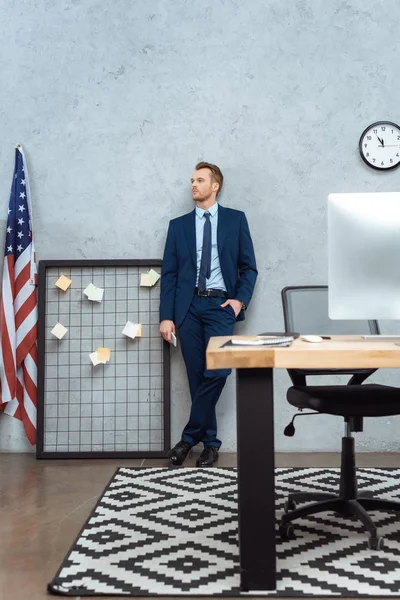 Избирательный фокус бизнесмена в костюме, стоящего со смартфоном возле американского флага за стеной в современном офисе — стоковое фото