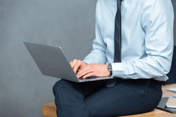 Обрезанный образ бизнесмена, сидящего на столе и работающего на ноутбуке в современном офисе — стоковое фото