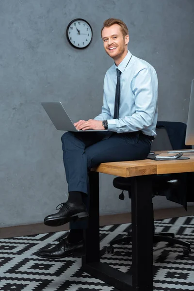 Сміється бізнесмен, сидить на столі і працює на ноутбуці в сучасному офісі — стокове фото
