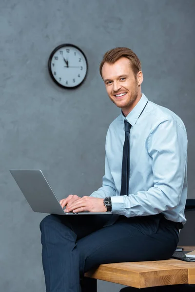 Happy young businessman sitting on table and working on laptop in modern office — Stock Photo
