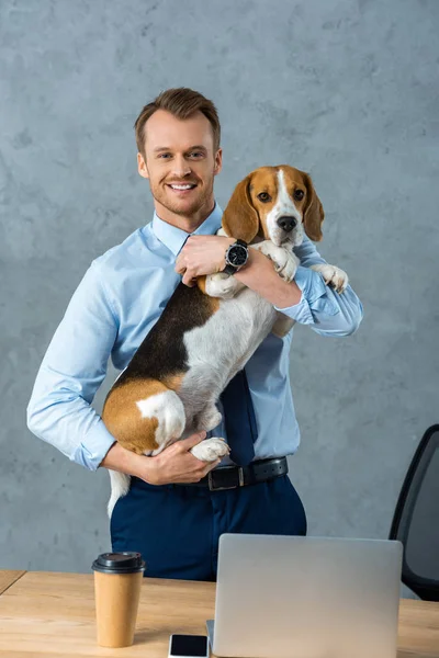 Joyeux jeune homme d'affaires tenant chien près de la table avec smartphone et ordinateur portable dans le bureau moderne — Photo de stock