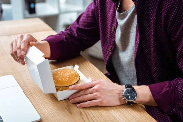 Частковий вид на бізнесмена, обідаючи з бургер за столом в сучасному офісі — стокове фото