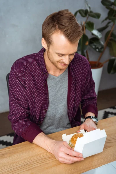 Visão de alto ângulo de homem de negócios feliz almoçando com hambúrguer à mesa no escritório moderno — Fotografia de Stock