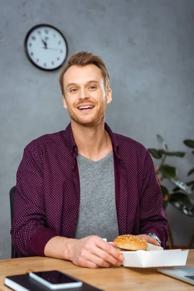 Jovem empresário alegre almoçando com hambúrguer à mesa no escritório moderno — Fotografia de Stock