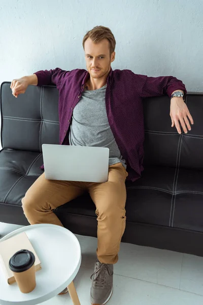 Heureux mâle pigiste travaillant sur ordinateur portable sur canapé dans le bureau à la maison — Photo de stock