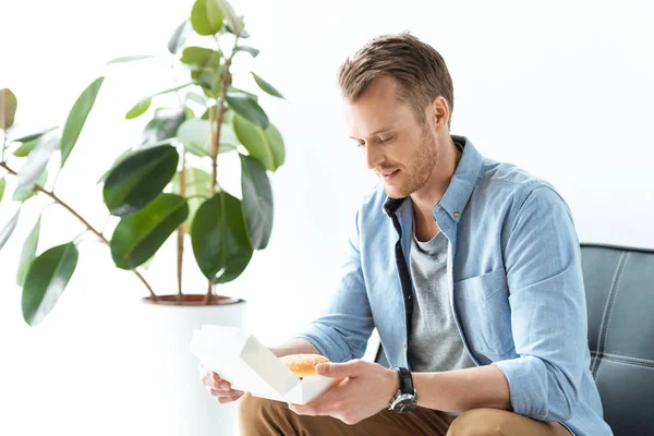 Cheerful young businessman having lunch with burger while sitting on sofa in office — Stock Photo