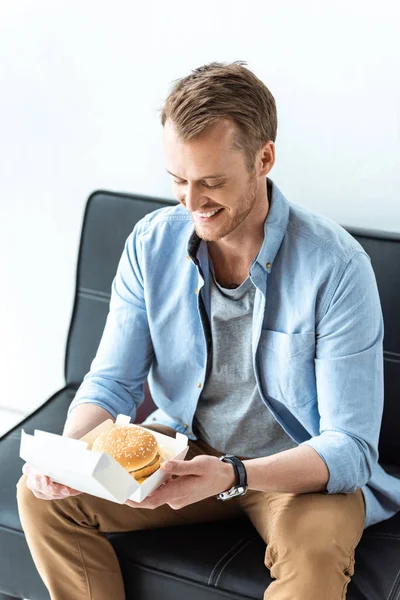 Feliz joven hombre de negocios almorzando con hamburguesa mientras está sentado en el sofá en la oficina - foto de stock