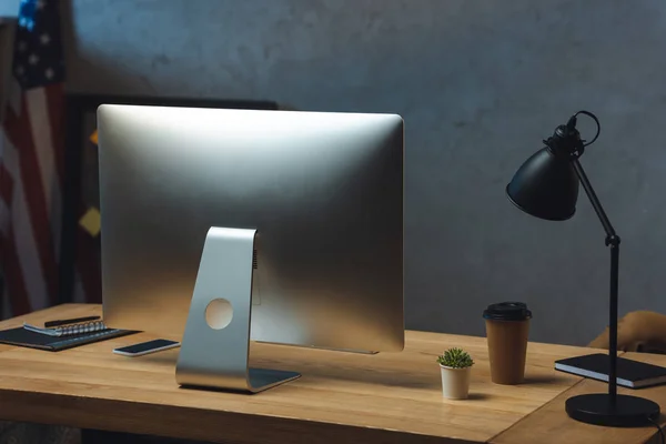 Enfoque selectivo de la mesa de trabajo con taza de café desechable, monitor de computadora y lámpara de escritorio en la oficina moderna - foto de stock