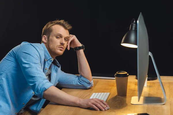 Jovem empresário sobrecarregado sentado à mesa com computador e xícara de café descartável durante a noite no escritório moderno — Fotografia de Stock