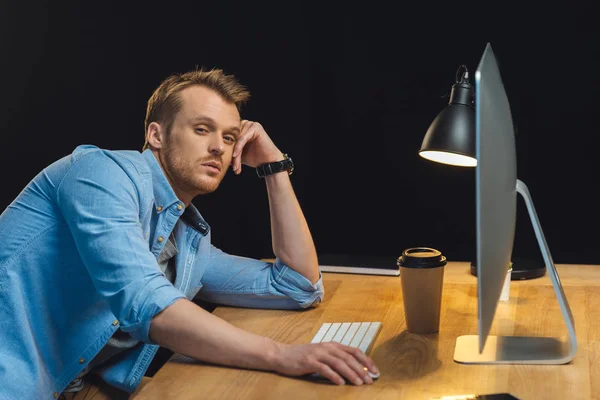Overworked young businessman sitting at table with computer and disposable coffee cup during late night in modern office — Stock Photo