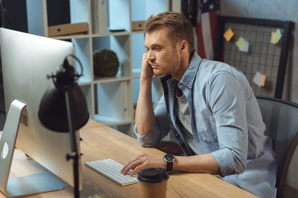 Enfoque selectivo de hombre de negocios sobrecargado de trabajo sentado en la mesa con el ordenador y la taza de café desechable durante la noche en la oficina moderna - foto de stock