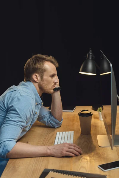 Handsome overworked businessman sitting at table with computer and disposable coffee cup under desk lump during late night in modern office — Stock Photo