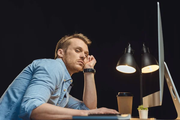 Blick aus dem niedrigen Winkel auf einen überlasteten jungen Geschäftsmann, der mit Computer und Einweg-Kaffeetasse unter dem Schreibtischklumpen in der späten Nacht im modernen Büro sitzt — Stockfoto
