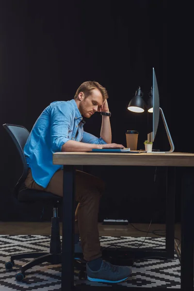 Junger überarbeiteter Geschäftsmann schläft am Tisch mit Schreibtischlampe, Kaffeetasse und Computer im Büro — Stockfoto