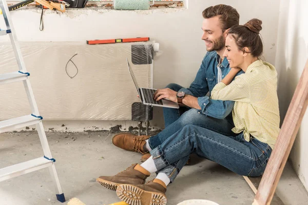 Vista lateral de la feliz pareja joven usando el ordenador portátil mientras está sentado en el suelo en la nueva casa - foto de stock