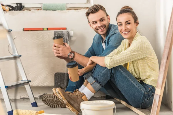 Glückliches junges Paar hält Pappbecher in der Hand und lächelt während der Reparatur in die Kamera — Stockfoto