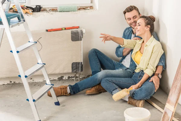 Feliz joven pareja sentada en el suelo y mirando hacia otro lado durante la mejora del hogar - foto de stock