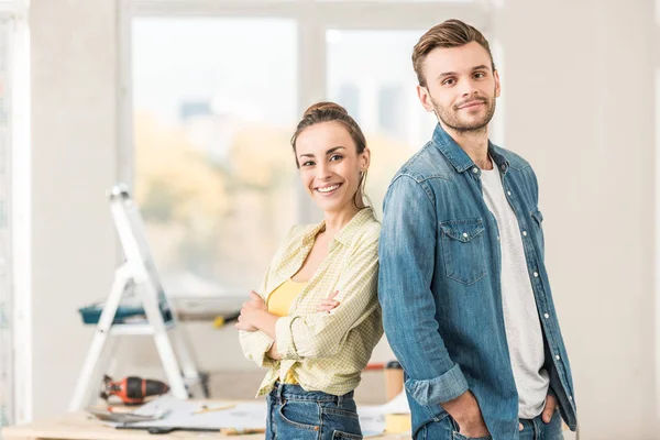 Heureux jeune couple debout dos à dos et souriant à la caméra dans la nouvelle maison — Photo de stock