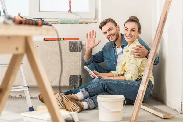 Feliz jovem casal usando smartphone e sorrindo para a câmera enquanto sentado no chão em nova casa — Fotografia de Stock