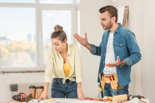 Emotional young man with toolbelt gesturing and quarreling with girlfriend during house repair — Stock Photo