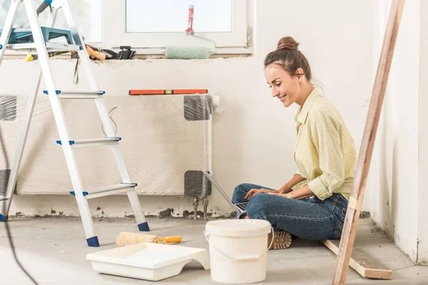 Vue latérale de la jeune femme souriante à l'aide d'un ordinateur portable tout en étant assis sur le sol à la nouvelle maison — Photo de stock