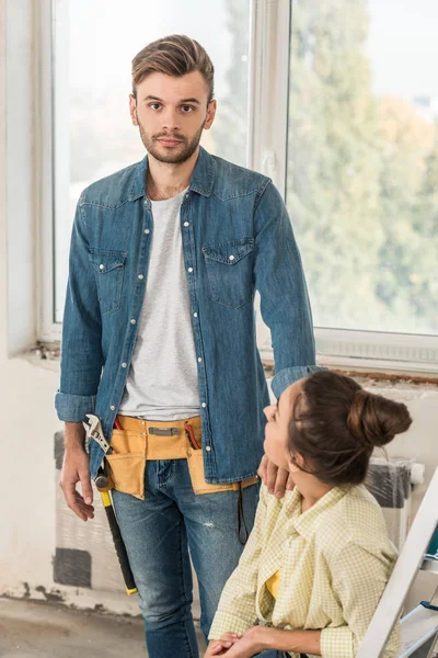Jeune homme avec ceinture d'outils regardant la caméra tout en réparant appartement avec petite amie — Photo de stock