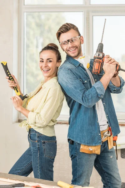 Feliz joven pareja sosteniendo martillo y taladro eléctrico durante la reparación de la casa - foto de stock