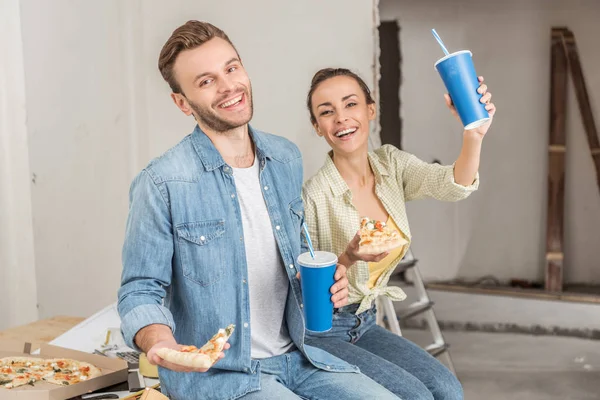 Happy young couple holding paper cups with drinking straws and pizza slices during repairment in new house — Stock Photo