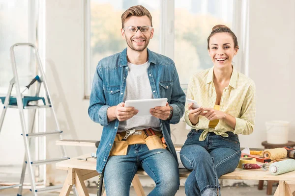 Happy young couple using digital devices and smiling at camera during renovation — Stock Photo